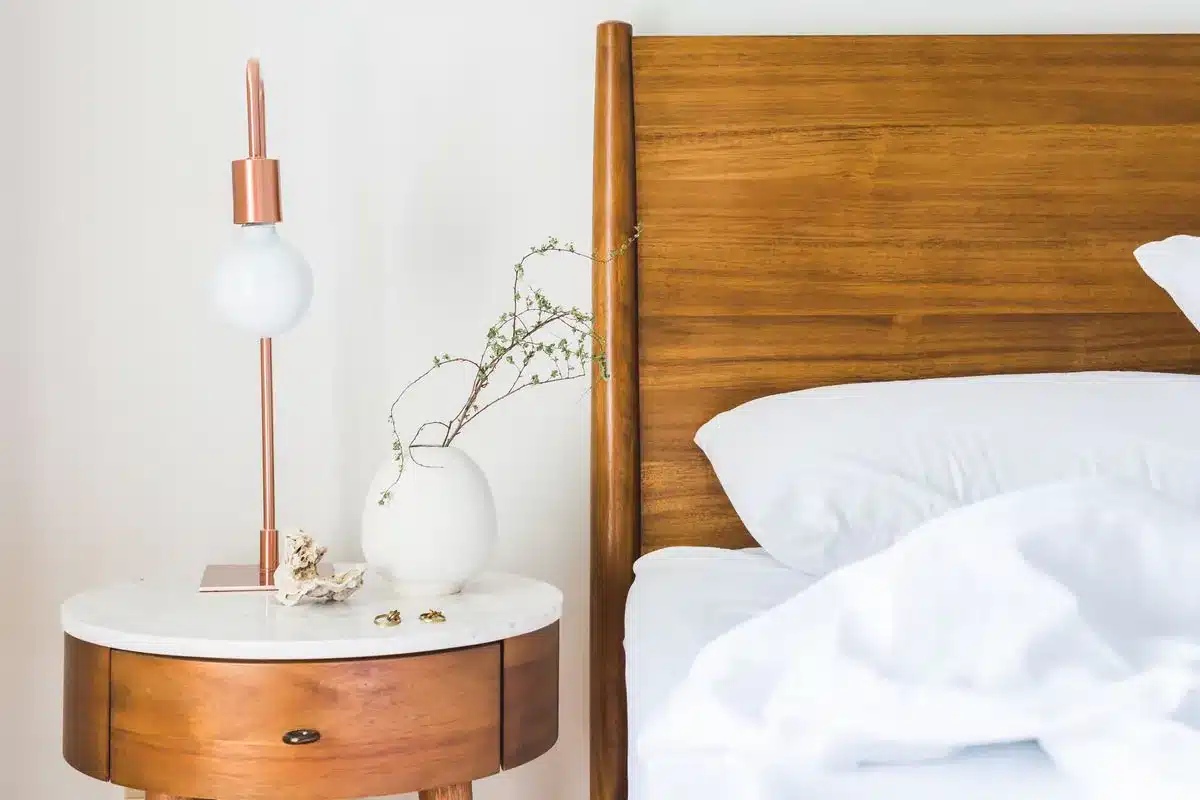 Criado-mudo redondo de tampo de mármore com abajur de cobre e vaso branco com um galho fino, ao lado de uma cama de cabeceira de madeira